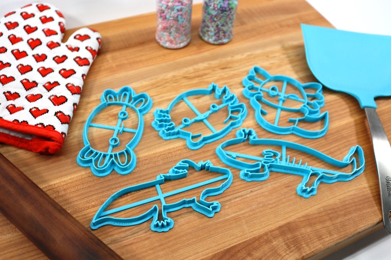 May include: Five blue plastic cookie cutters shaped like axolotls, a turtle, and a hedgehog on a wooden cutting board. There are two jars of sprinkles in the background.