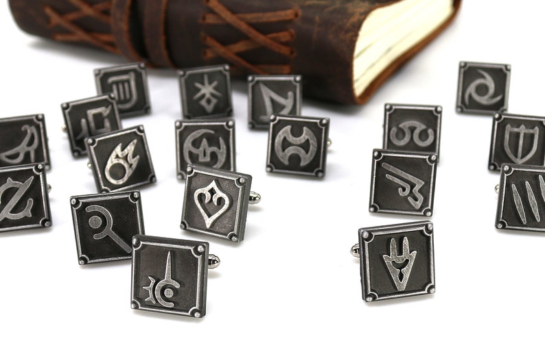 May include: A set of 16 silver cufflinks featuring various symbols, each with a black background and a silver design. The cufflinks are arranged on a white surface.