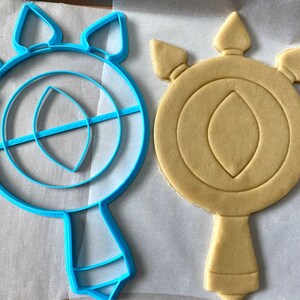 May include: A blue plastic cookie cutter and a light brown sugar cookie, both shaped like a stylized eye with a teardrop-shaped pupil and three points at the top.