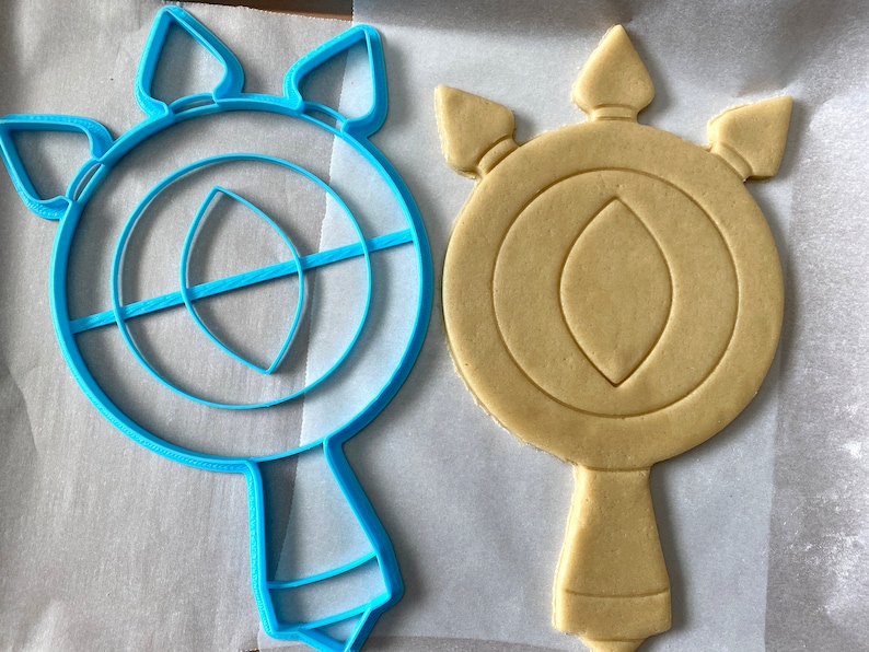 May include: A blue plastic cookie cutter and a light brown sugar cookie, both shaped like a stylized eye with a teardrop-shaped pupil and three points at the top.