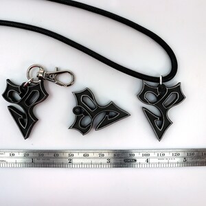May include: Three black metal pendants with a geometric design, one attached to a black cord necklace and another to a silver key chain. A ruler is included for scale.