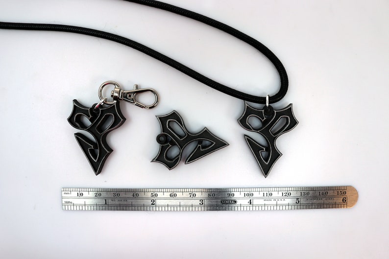 May include: Three black metal pendants with a geometric design, one attached to a black cord necklace and another to a silver key chain. A ruler is included for scale.