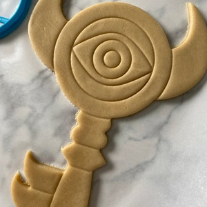 Op de afbeelding: Een lichtbruine koek in de vorm van een sleutel met een groot oogontwerp in het midden. De koek ligt op een wit marmeren oppervlak.