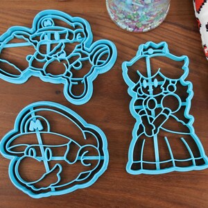 May include: Three blue plastic cookie cutters shaped like characters from the video game Super Mario Bros. The cutters are on a wooden surface. The characters are Mario, Luigi, and Princess Peach.