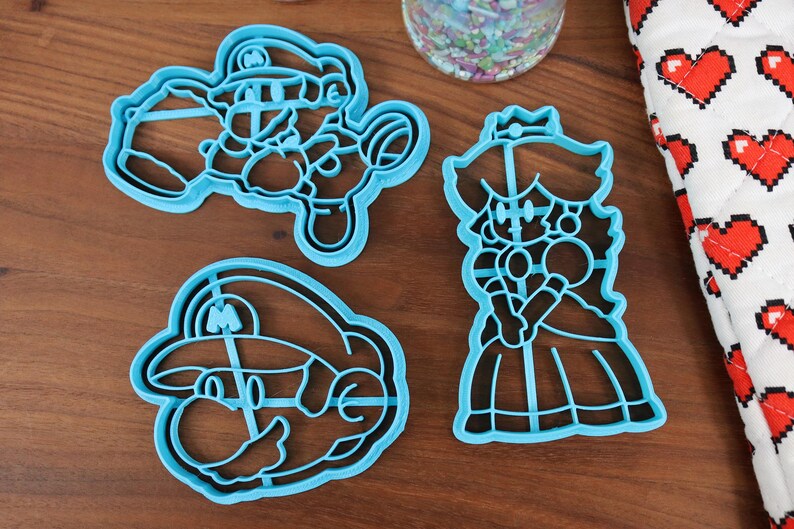 May include: Three blue plastic cookie cutters shaped like characters from the video game Super Mario Bros. The cutters are on a wooden surface. The characters are Mario, Luigi, and Princess Peach.