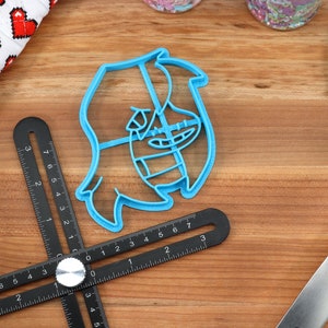 May include: A blue plastic cookie cutter shaped like a cartoon character with a wide smile and a large nose. The cutter is on a wooden surface with a ruler and a spatula.