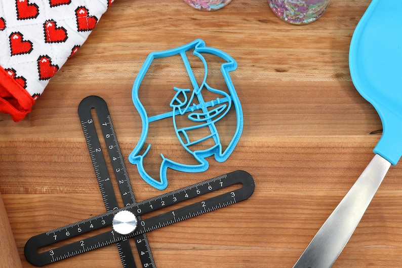 May include: A blue plastic cookie cutter shaped like a cartoon character with a wide smile and a large nose. The cutter is on a wooden surface with a ruler and a spatula.