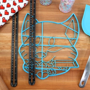 May include: A blue plastic cookie cutter shaped like a dog's face, a black metal ruler, a wooden rolling pin, a red and white oven mitt, a blue spatula with a silver handle, and two jars of sprinkles on a wooden surface.
