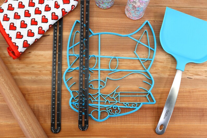 May include: A blue plastic cookie cutter shaped like a dog's face, a black metal ruler, a wooden rolling pin, a red and white oven mitt, a blue spatula with a silver handle, and two jars of sprinkles on a wooden surface.