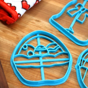 May include: A set of three blue plastic cookie cutters shaped like an Easter egg, a bunny, and a dress. The cutters are on a wooden surface.
