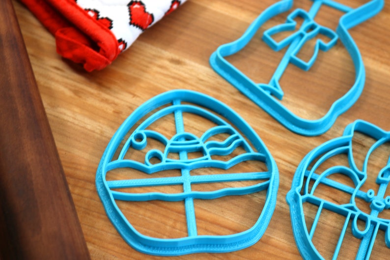 May include: A set of three blue plastic cookie cutters shaped like an Easter egg, a bunny, and a dress. The cutters are on a wooden surface.