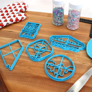 May include: A set of blue plastic cookie cutters on a wooden surface. The cutters are shaped like a house, an alien, a sign that says "Friz Aegvz Mellowwe", a circle, and a square. There are also two jars of sprinkles, a rolling pin, a spatula, and a red oven mitt with white hearts.