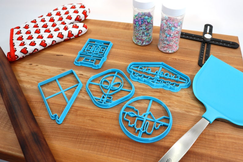 May include: A set of blue plastic cookie cutters on a wooden surface. The cutters are shaped like a house, an alien, a sign that says "Friz Aegvz Mellowwe", a circle, and a square. There are also two jars of sprinkles, a rolling pin, a spatula, and a red oven mitt with white hearts.