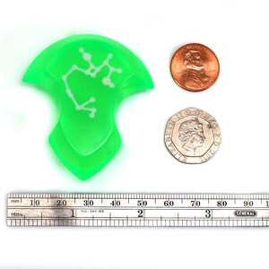 May include: A green plastic pick with a white constellation design. The pick is about 5 cm long and is next to a ruler and two coins for scale.