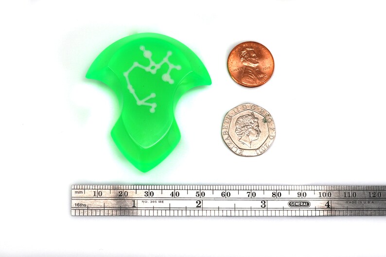 May include: A green plastic pick with a white constellation design. The pick is about 5 cm long and is next to a ruler and two coins for scale.