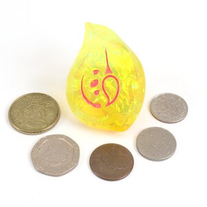 May include: A yellow teardrop-shaped resin dice with a red symbol on it. The dice is surrounded by six silver coins.