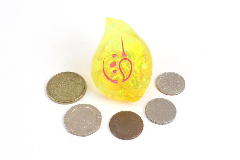 May include: A yellow teardrop-shaped resin dice with a red symbol on it. The dice is surrounded by six silver coins.