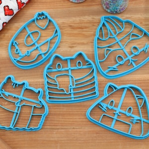 May include: Five blue plastic cookie cutters shaped like cats. The cutters are on a wooden surface.