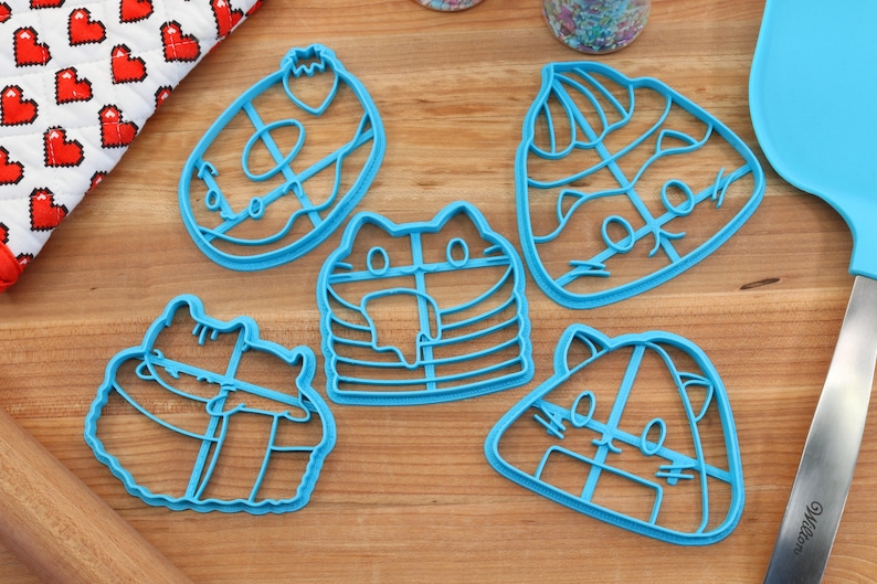 May include: Five blue plastic cookie cutters shaped like cats. The cutters are on a wooden surface.