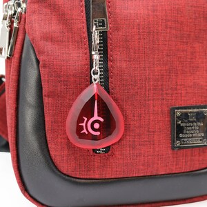 May include: A red crossbody bag with a black zipper and a black leather trim. The bag has a silver zipper pull with a red teardrop-shaped charm with a black symbol. The bag has a silver tag with the text "Where is the heart to receive good where" and the text "L.C.WOMAN".