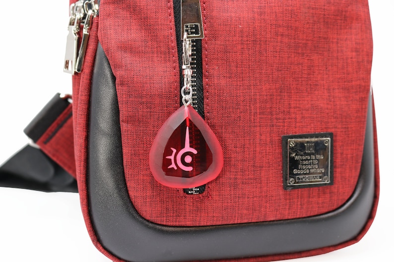 May include: A red crossbody bag with a black zipper and a black leather trim. The bag has a silver zipper pull with a red teardrop-shaped charm with a black symbol. The bag has a silver tag with the text "Where is the heart to receive good where" and the text "L.C.WOMAN".