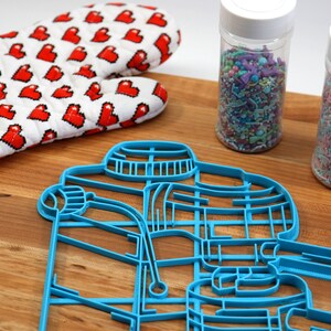 May include: A blue plastic cookie cutter shaped like a coffee maker, two jars of colorful sprinkles, a red heart-patterned oven mitt, and a black ruler on a wooden surface.