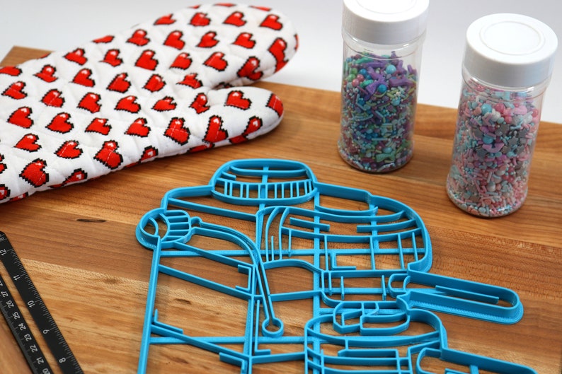 May include: A blue plastic cookie cutter shaped like a coffee maker, two jars of colorful sprinkles, a red heart-patterned oven mitt, and a black ruler on a wooden surface.