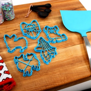 May include: A set of blue plastic cookie cutters shaped like different dog breeds, including a papillon, a poodle, and a terrier. The cutters are on a wooden cutting board with a blue spatula and a red and white oven mitt.