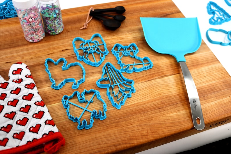 May include: A set of blue plastic cookie cutters shaped like different dog breeds, including a papillon, a poodle, and a terrier. The cutters are on a wooden cutting board with a blue spatula and a red and white oven mitt.
