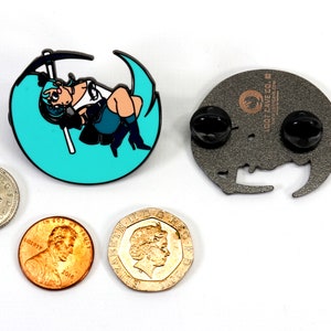May include: A teal blue enamel pin featuring a cartoon character holding a scythe, sitting on a crescent moon. The back of the pin is silver with a black rubber clutch and the text "Loot Cave Co."