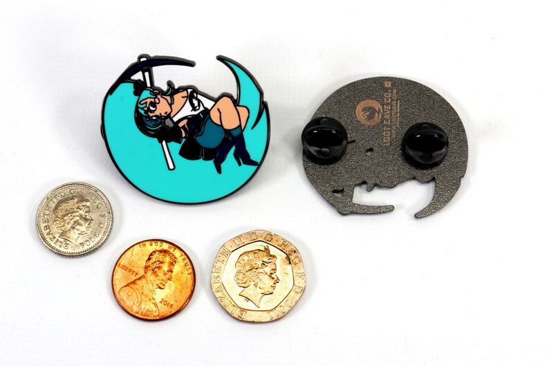 May include: A teal blue enamel pin featuring a cartoon character holding a scythe, sitting on a crescent moon. The back of the pin is silver with a black rubber clutch and the text "Loot Cave Co."