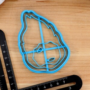 May include: A blue plastic cookie cutter shaped like a face with a double outline. The cutter is on a wooden surface.
