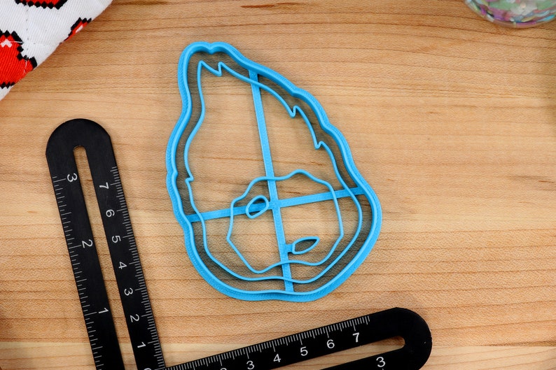 May include: A blue plastic cookie cutter shaped like a face with a double outline. The cutter is on a wooden surface.