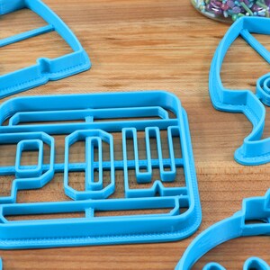 May include: A set of blue plastic cookie cutters in various shapes, including a rocket, a car, and the word "POW!"