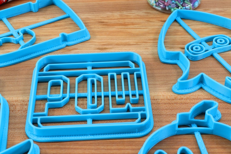 May include: A set of blue plastic cookie cutters in various shapes, including a rocket, a car, and the word "POW!"