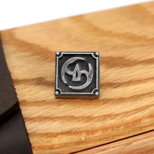 May include: A silver metal square with a stylized symbol inside. The symbol is a circle with three curved lines inside. The square has a wood grain background.