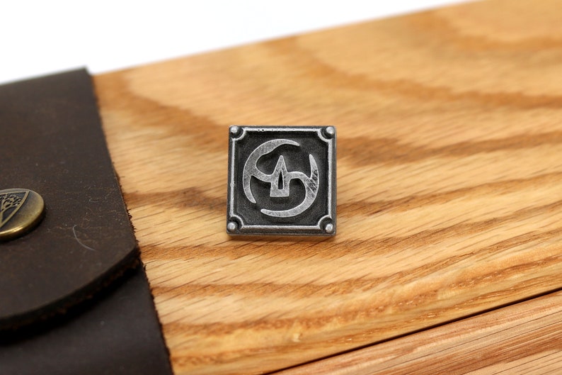 May include: A silver metal square with a stylized symbol inside. The symbol is a circle with three curved lines inside. The square has a wood grain background.