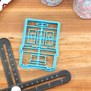 May include: A blue plastic cookie cutter shaped like a slot machine. The cookie cutter has three windows with a line in each window. The cookie cutter is on a wooden surface.