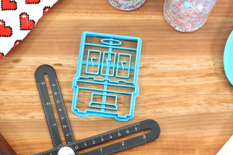 May include: A blue plastic cookie cutter shaped like a slot machine. The cookie cutter has three windows with a line in each window. The cookie cutter is on a wooden surface.
