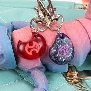 Puede incluir: Dos llaveros con anillos de metal plateado. Un charm es rojo con un s&iacute;mbolo morado, el otro es morado con un s&iacute;mbolo plateado y purpurina.
