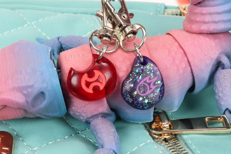 Puede incluir: Dos llaveros con anillos de metal plateado. Un charm es rojo con un s&iacute;mbolo morado, el otro es morado con un s&iacute;mbolo plateado y purpurina.