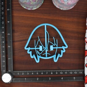 May include: A light blue plastic cookie cutter shaped like a cartoon character with short, spiky hair and a serious expression.