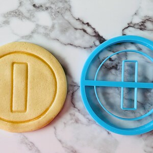 May include: A blue plastic cookie cutter shaped like a gold coin with a vertical line in the center. Next to it is a cookie that has been cut with the cookie cutter.