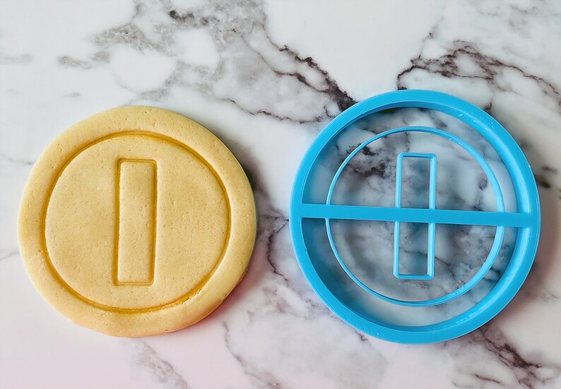 May include: A blue plastic cookie cutter shaped like a gold coin with a vertical line in the center. Next to it is a cookie that has been cut with the cookie cutter.