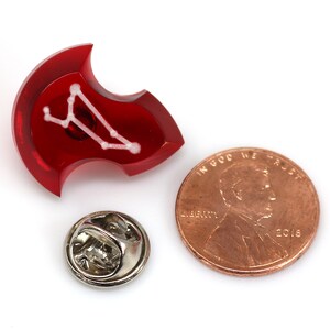 May include: Red enamel pin with a white constellation design. The pin is shaped like a shield and has a silver pin back.