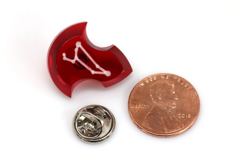 May include: Red enamel pin with a white constellation design. The pin is shaped like a shield and has a silver pin back.