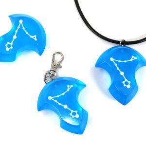 K&ouml;nnte beinhalten: Drei blaue Harz-Anh&auml;nger mit einem wei&szlig;en Sternbild-Design. Ein Anh&auml;nger ist an einer schwarzen Schnurkette, einer an einem silbernen Schl&uuml;sselanh&auml;nger und einer auf einer wei&szlig;en Oberfl&auml;che.