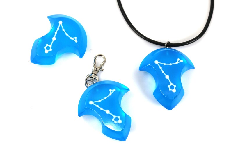 K&ouml;nnte beinhalten: Drei blaue Harz-Anh&auml;nger mit einem wei&szlig;en Sternbild-Design. Ein Anh&auml;nger ist an einer schwarzen Schnurkette, einer an einem silbernen Schl&uuml;sselanh&auml;nger und einer auf einer wei&szlig;en Oberfl&auml;che.