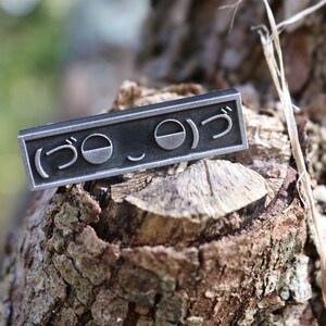 May include: A silver metal pin with the Japanese characters "(うo_o)つ" engraved on it. The pin is sitting on a piece of tree bark.