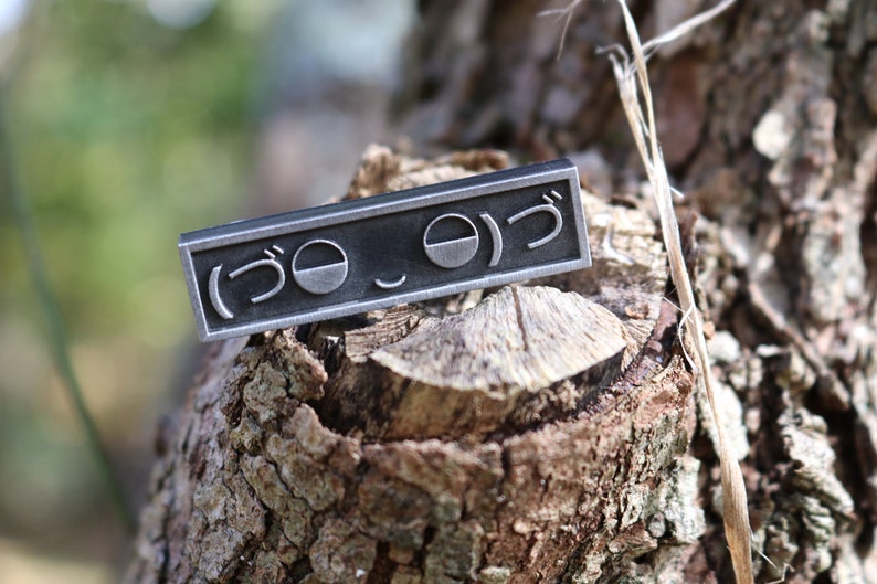May include: A silver metal pin with the Japanese characters "(うo_o)つ" engraved on it. The pin is sitting on a piece of tree bark.
